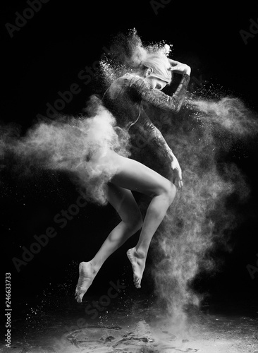 Beautiful slim girl wearing a gymnastic bodysuit covered with clouds of the flying white powder jumps dancing on a dark. Artistic conceptual and advertising black and white photo.