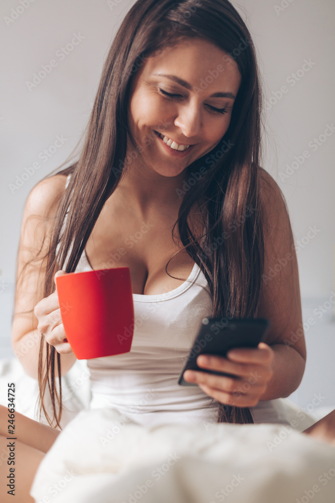 Young smiling woman drinking coffee at home in her bed and checking her smartphone Young smiling woman drinking coffee at home in her bed and checking her smartphone