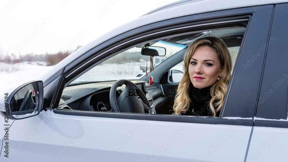 Blonde woman sits at a white car steering wheel and staring through the open window.