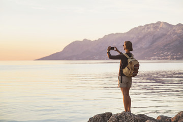 Young woman using smart phone taking photo of the sea at sunset, travel and active lifestyle concept