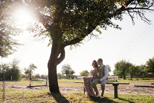 Wedding at sunset. Couple is sitting on a bench under a tree. Beige dress with sparkles. Light suit with a bow tie. The bride and groom embrace and kiss. © malysheva