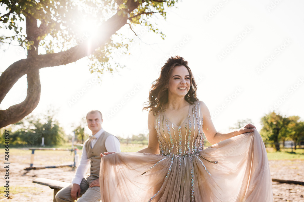 Wedding at sunset. Couple is sitting on a bench under a tree. Beige dress with sparkles. Light suit with a bow tie. The bride and groom embrace and kiss.