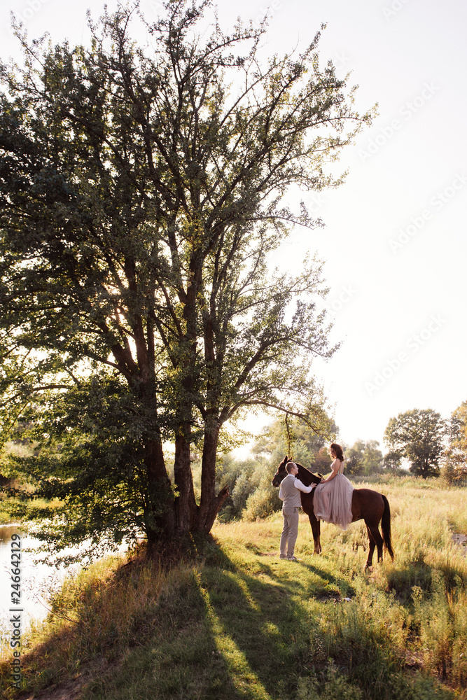 Wedding at sunset. Couple is sitting on a bench under a tree. Beige dress with sparkles. Light suit with a bow tie. The bride and groom embrace and kiss.