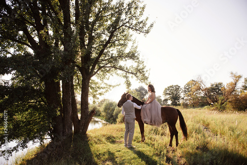 Wedding at sunset. Couple is sitting on a bench under a tree. Beige dress with sparkles. Light suit with a bow tie. The bride and groom embrace and kiss.