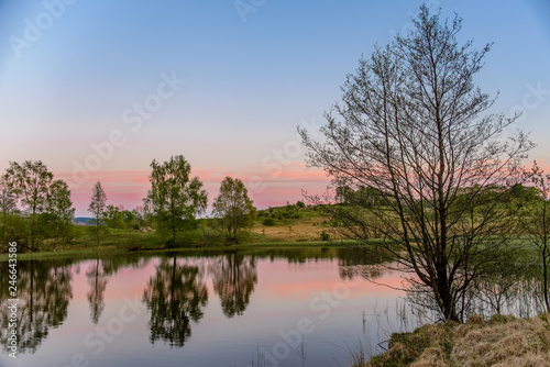 A tranquil sunset scene with a pink and blue sky and trees reflected in a calm lake.