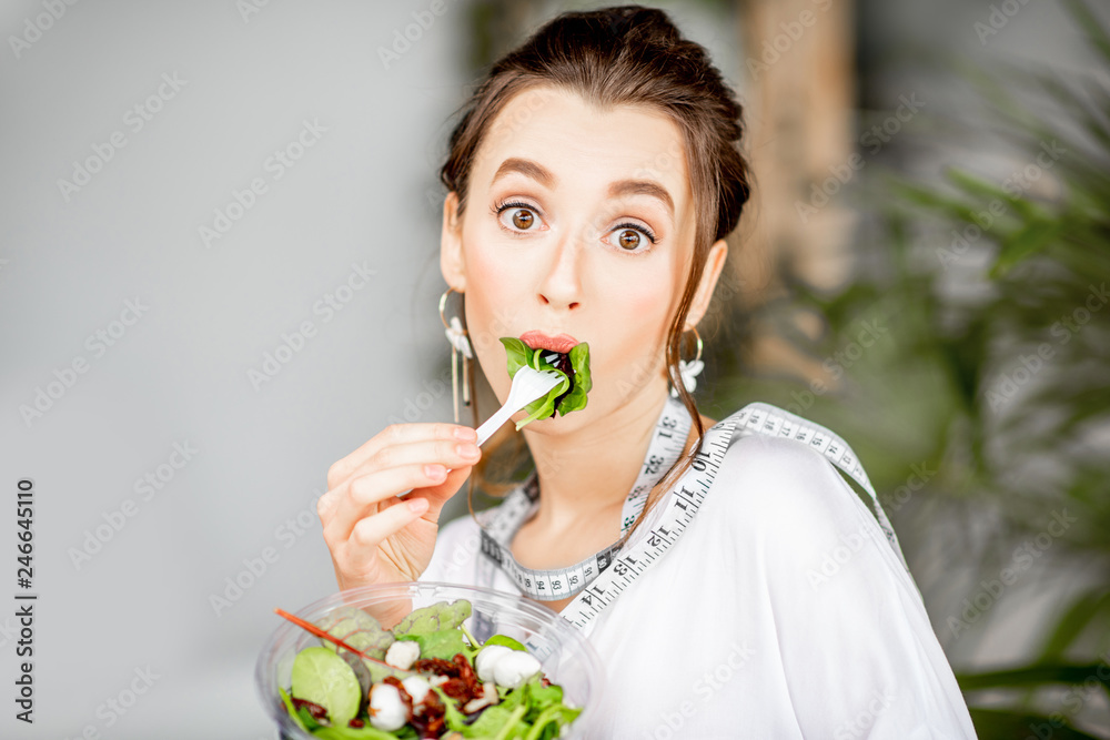 Young and beautiful woman in white shirt eating healthy salad indoors with green plants on the background. Healthy eating and beauty concept Young and beautiful woman in white shirt eating healthy salad indoors with green plants on the background. Healthy eating and beauty concept