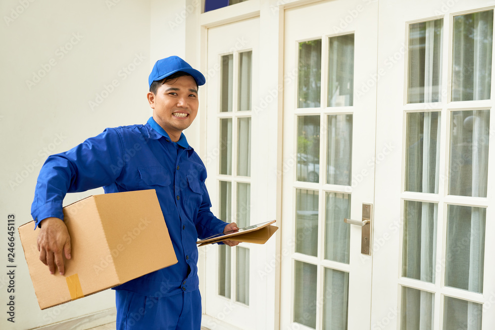 Happy courier in uniform holding big packages when standing at entrance of house Happy courier in uniform holding big packages when standing at entrance of house
