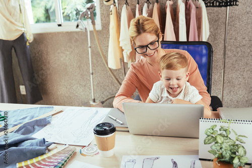 Cheerful female fashion designer working on laptop with son on her laps