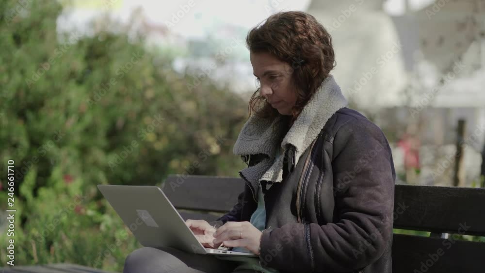 Focused curly female freelancer working with laptop outdoor. Thoughtful mature lady typing on laptop while sitting on wooden bench during warm spring day. Technology concept