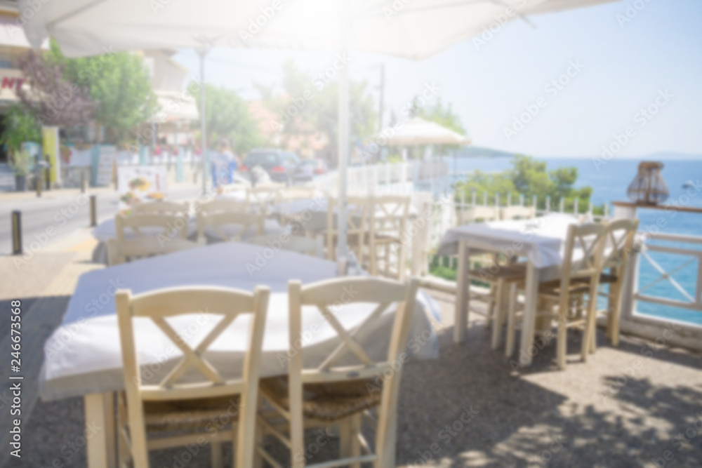 Obraz premium Restaurant tables and chairs below the sunshade on the terrace on the seashore. Blurred scene of the restaurant in Neos marmaras, Greece.