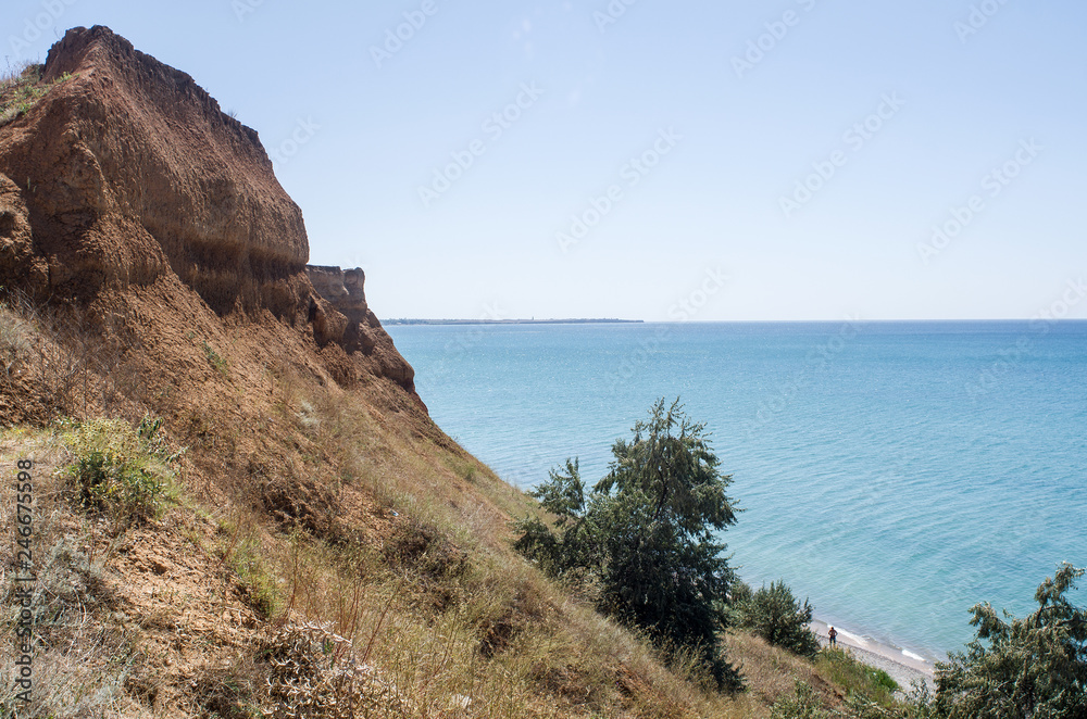 High cliff on the beach, in the summer afternoon, with greenery on the slope, from which far visible sea space with the coastline on the horizon, copy space.