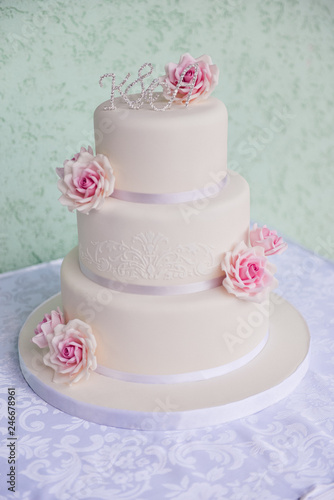 White three-tiered wedding cake with pink roses made of mastic stands on the table a delicate green background. letters bride and groom