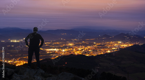 A man stands on top of a mountain, against the backdrop of the lights of the night city. © poliki