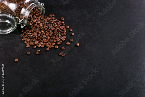 Fragrant coffee beans are scattered from a jar on a rustic tabletop background. Banner copy space.