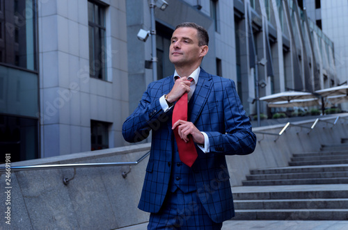 Charismatic man in a plaid suit on the background of the business center (office). Business men in a suit straightens (removes) a red tie. Freedom, the will of office work, vacation, weekend