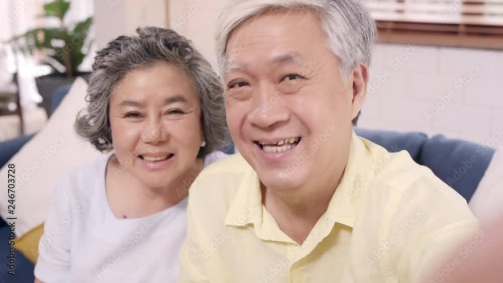 Asian elderly couple using smartphone video conference with grandchild while lying on sofa in living room at home. Enjoying time lifestyle senior family at home concept. Portrait looking at camera. Asian elderly couple using smartphone video conference with grandchild while lying on sofa in living room at home. Enjoying time lifestyle senior family at home concept. Portrait looking at camera.