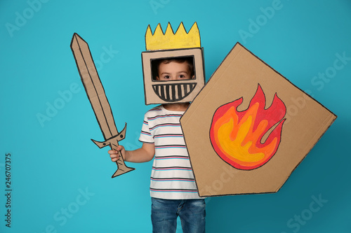 Cute little child in cardboard armor on color background