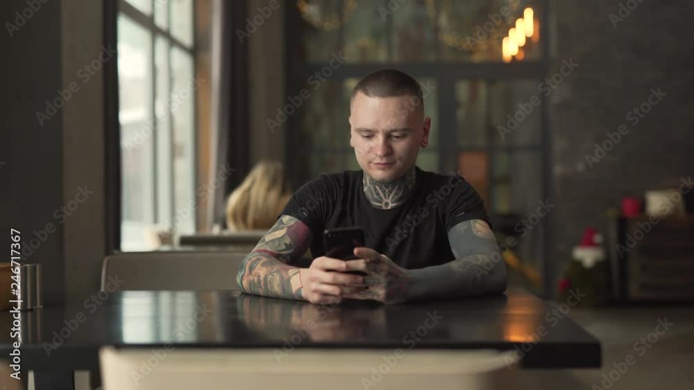 Young charming man with tattoos on his neck and arms is sitting in a cafe indoors and typing a message on a phone. Man is smiling and looking pleased.
