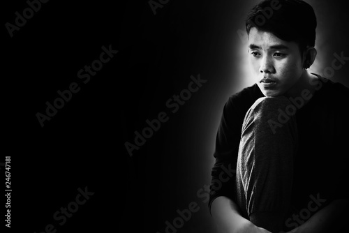 Boy with lower head and arm around knee sitting on corner of room in the dark