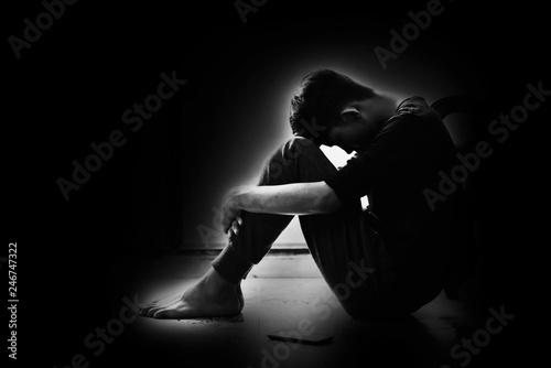 Boy with lower head and arm around knee sitting on corner of room in the dark