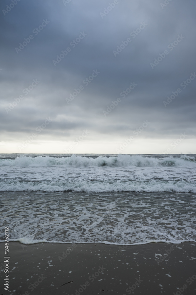 Sunset on horizon, ocean waves and sand coastline in front, gray cloudy sky