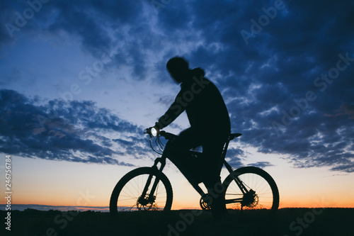 Silhouette of a young man in a trip on the mountain bike with head lamp in a dark conditions against a cloudy evening sky . Low angle view with motion blur