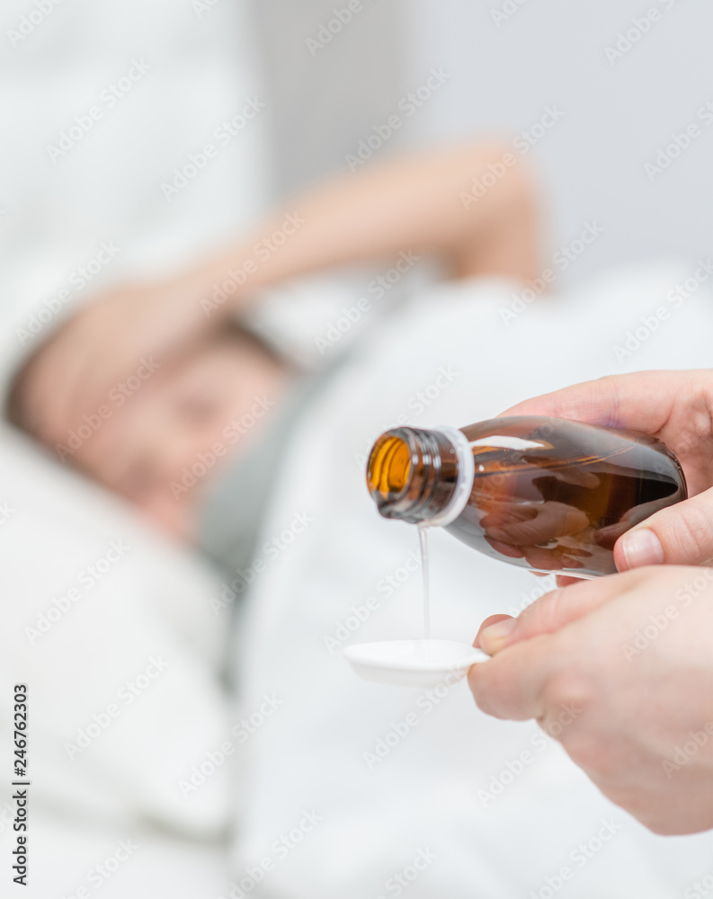 Sick little boy awaits his medication pouring in a spoon in the foreground Sick little boy awaits his medication pouring in a spoon in the foreground
