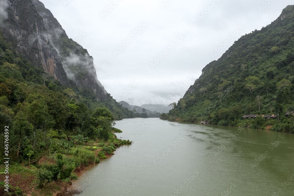 Nam Ou river view with small long boat sailing. A valley surrounded by steep limestone cliffs covered with jungle. Image taken in Nong Khiaw - a village in the Luang Prabang Province of northern Laos. Nam Ou river view with small long boat sailing. A valley surrounded by steep limestone cliffs covered with jungle. Image taken in Nong Khiaw - a village in the Luang Prabang Province of northern Laos.