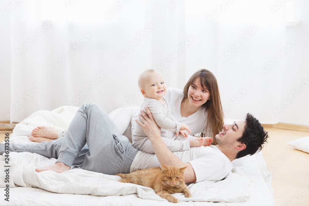 The family spends together time - beautiful mommy charming dad with cute one year old baby play with ginger cat in white home interior