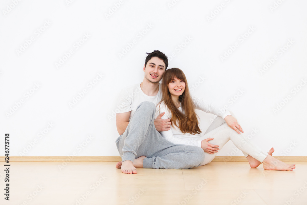 Happy young family at home. Beautiful young couple in white pajamas kisses and hugs sitting on the floor during weekends at home. Love concept, white background Happy young family at home. Beautiful young couple in white pajamas kisses and hugs sitting on the floor during weekends at home. Love concept, white background