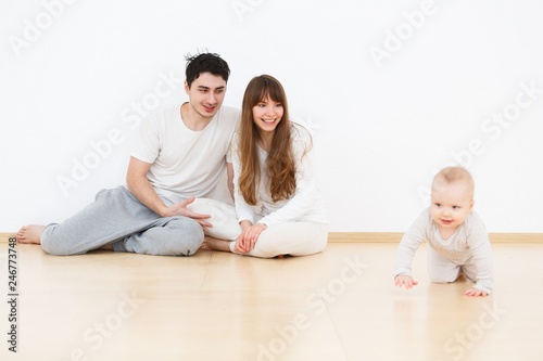 Positive parents call their little cute baby to play sitting on the floor in white pajamas on sunny sunday at home. Warm real scene. White wall background