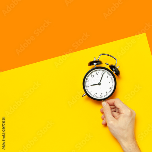 Flat lay top view male hand holding black vintage alarm clock on bright yellow orange color background with copy space. Creative Time concept minimal style, morning time to work