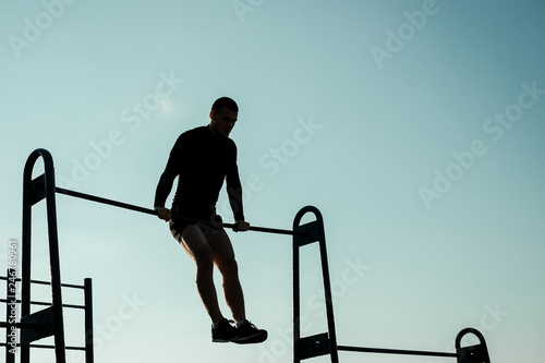 A young muscular man pulls himself up on a bar in the background of the sun on the street.
