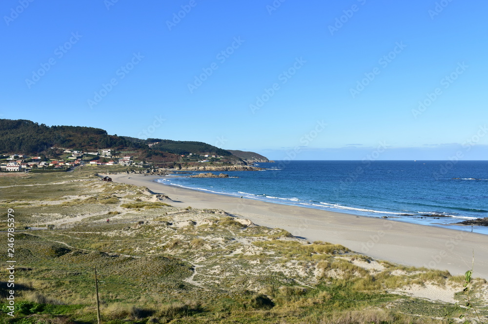 Beach and sand dunes with morning light. Rocks and blue sea with small waves and foam. Sunny day, clear sky. Galicia, Spain.