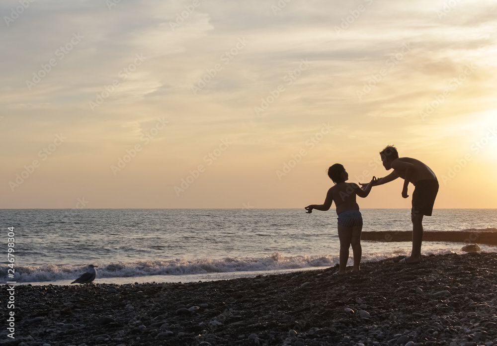 Silhouettes of children at sunset, feeding a seagull, against the background of the sea.