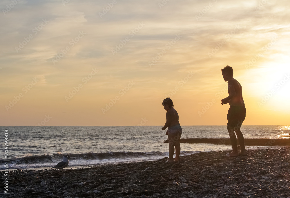 Silhouettes of children at sunset, feeding a seagull, against the background of the sea.