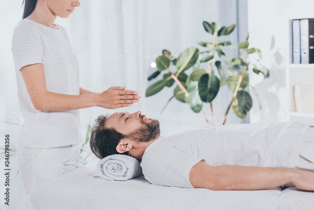 Obraz premium side view of calm bearded man with closed eyes receiving reiki treatment on massage table