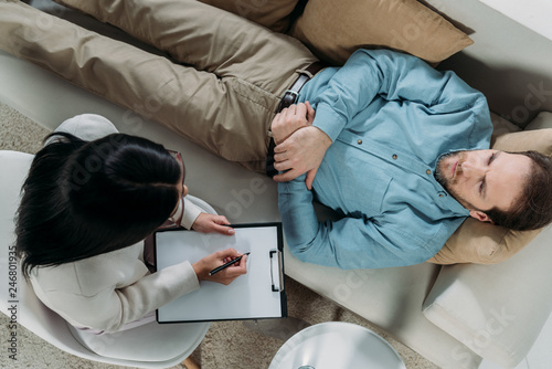 overhead view of psychotherapist writing on clipboard and male patient lying on couch
