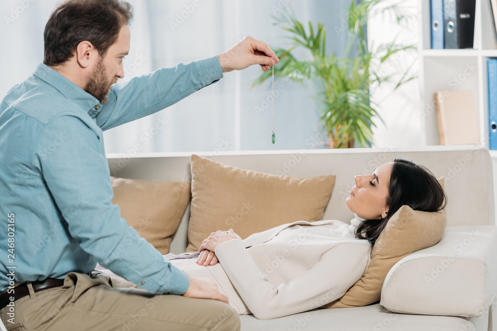 side view of bearded hypnotist holding pendulum and hypnotising young woman with closed eyes lying on couch side view of bearded hypnotist holding pendulum and hypnotising young woman with closed eyes lying on couch