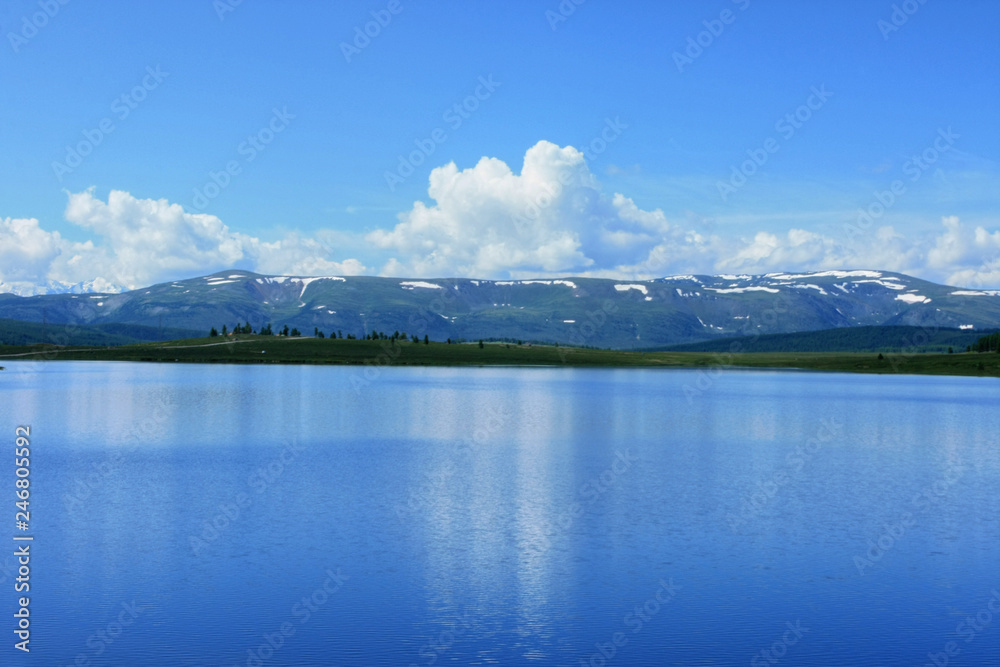 Beautiful sunny day overlooking the lake in the Altai Republic in Russia. White clouds in the blue sky reflected in the water and snow in the mountains far away on the horizon