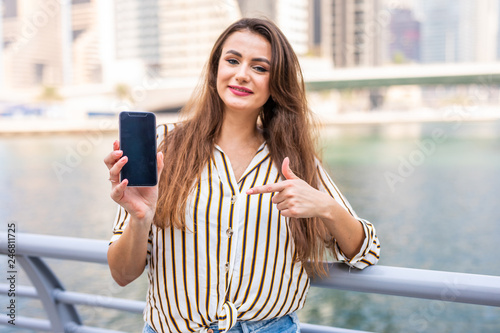 Young pretty woman pointed on the phone screen for advertising standing on Dubai marina promenade photo