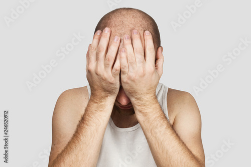 A bald man covers his face with his hands and cries from hopelessness, loneliness and problems on a gray background