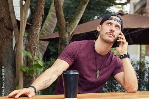 Close-up portrait of bored good-looking mixed race man with disposable coffee cup, wearing hat, holding cell phone, talking with friend looking up, having pensive expression. People, communication.