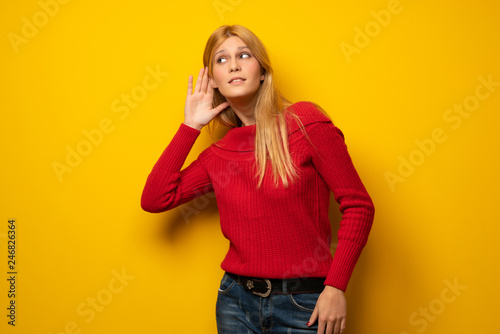 Blonde woman over yellow wall listening to something by putting hand on the ear