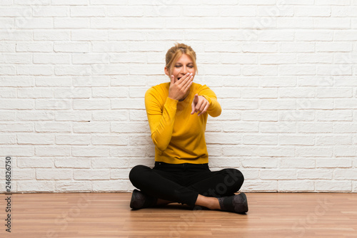 Young girl sitting on the floor pointing with finger at someone and laughing