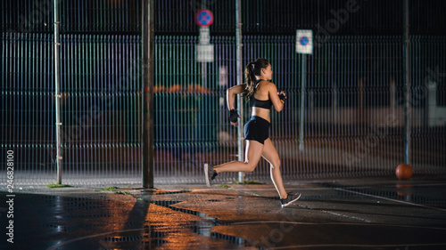 Beautiful Energetic Fitness Girl is Sprinting in a Fenced Outdoor Basketball Court. She's Running at Night After Rain in a Residential Neighborhood Area.