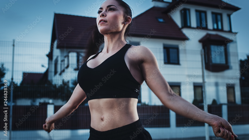 Close Up Shot of a Beautiful Busty Fitness Girl Skipping/Jumping Rope. She is Doing a Workout in a Fenced Outdoor Basketball Court. Evening Shot After Rain in a Residential Neighborhood Area.