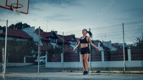 Beautiful Energetic Fitness Girl Skipping/Jumping Rope. She is Doing a Workout in a Fenced Outdoor Basketball Court. Evening Shot After Rain in a Residential Neighborhood Area.