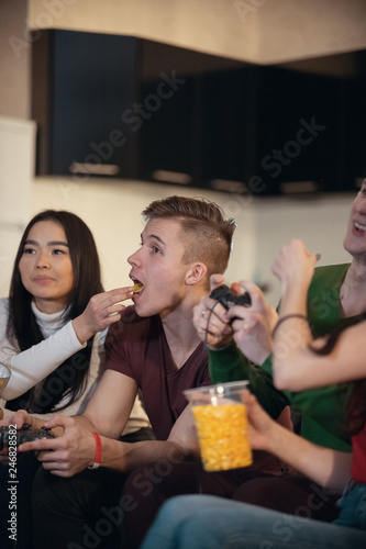 Company of young friends spending time together playing games and eating food. Young man opens his mouth and his girlfriend feed him