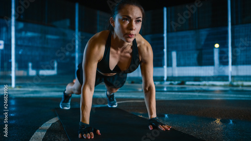 Beautiful Sporty Fitness Girl Doing Push Up Exercises. She is Doing a Workout in a Fenced Outdoor Basketball Court. Night Footage After Rain in a Residential Neighborhood Area. © Gorodenkoff